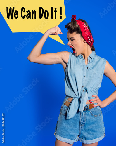 young woman in retro clothing showing muscles and shouting with we can do it speech bubble isolated on blue