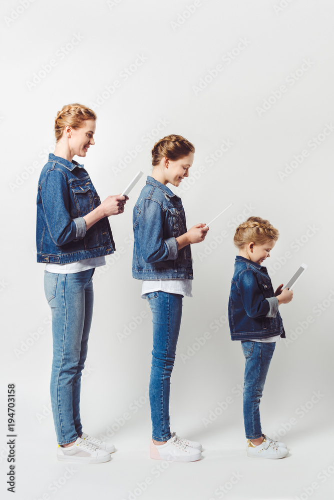 Fototapeta premium smiling mother and daughters in similar denim clothing standing in row and using tablets isolated on grey