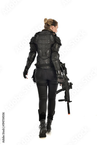 full length portrait of female soldier wearing black tactical armour, standing with back to the camera holding a gun, isolated on white studio background.