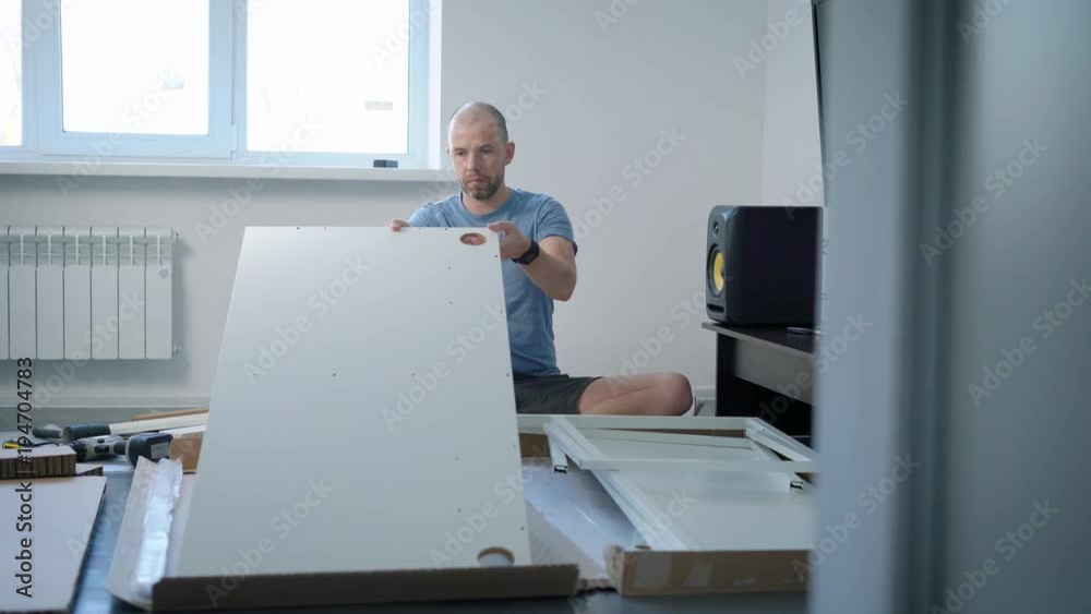 young man is sorting and exploring parts of new object of furniture, sitting on a floor in his clear flat young man is sorting and exploring parts of new object of furniture, sitting on a floor in his clear flat