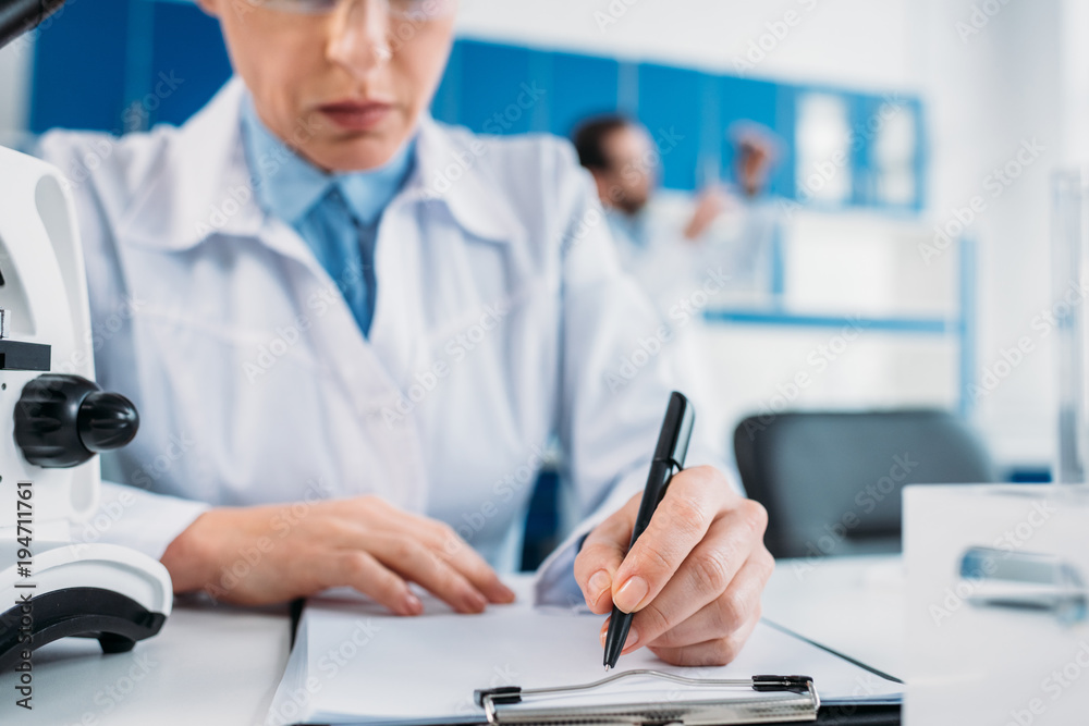 partial view of female scientist making notes in notepad at workplace with colleague behind in lab partial view of female scientist making notes in notepad at workplace with colleague behind in lab