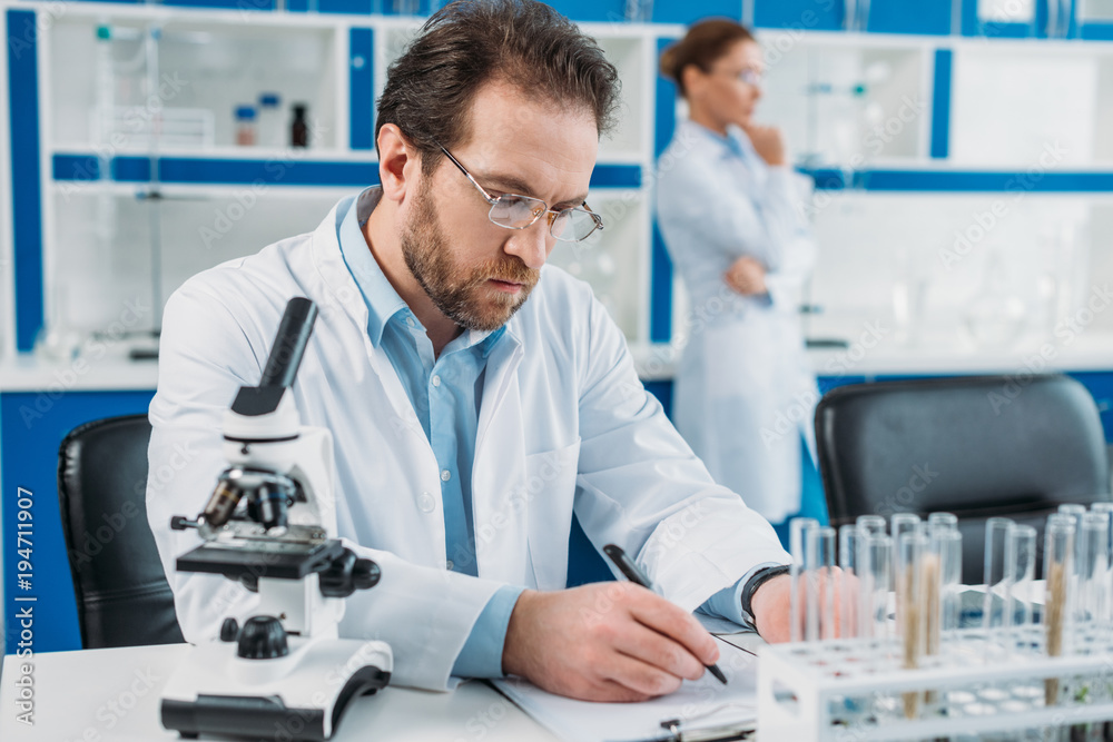 selective focus of scientist in white coat and eyeglasses making notes in notepad at workplace with microscope in laboratory selective focus of scientist in white coat and eyeglasses making notes in notepad at workplace with microscope in laboratory