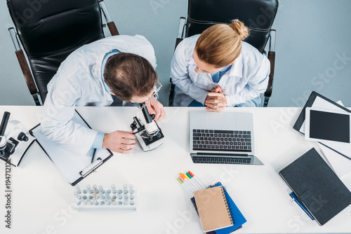 overhead view of scientific researchers in white coats working together at workplace with microscope and laptop in laboratory