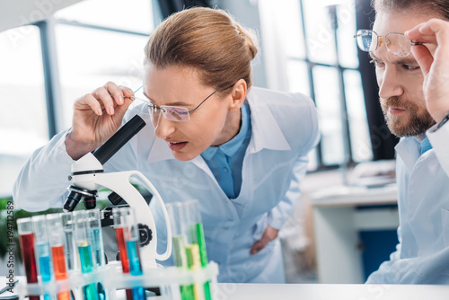 scientist in lab coat looking through microscope on reagent with colleague near by in laboratory