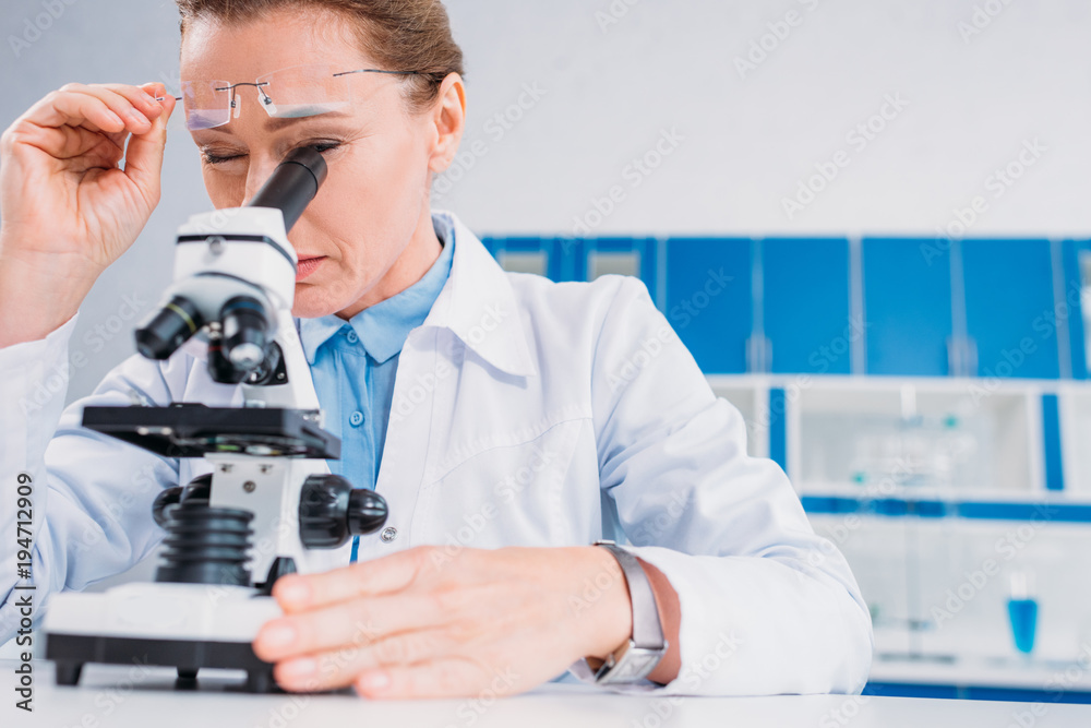 female scientist in lab coat and eyeglasses looking through microscope on reagent in lab female scientist in lab coat and eyeglasses looking through microscope on reagent in lab