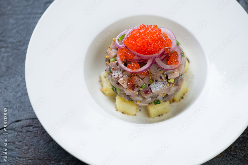 Herring fillet tartare on a bed of potato with red caviar topping served in a white plate, closeup, selective focus Herring fillet tartare on a bed of potato with red caviar topping served in a white plate, closeup, selective focus