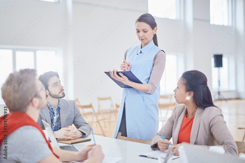 Young confident coach standing by desk and making notes in document or plan while three young managers listening to her