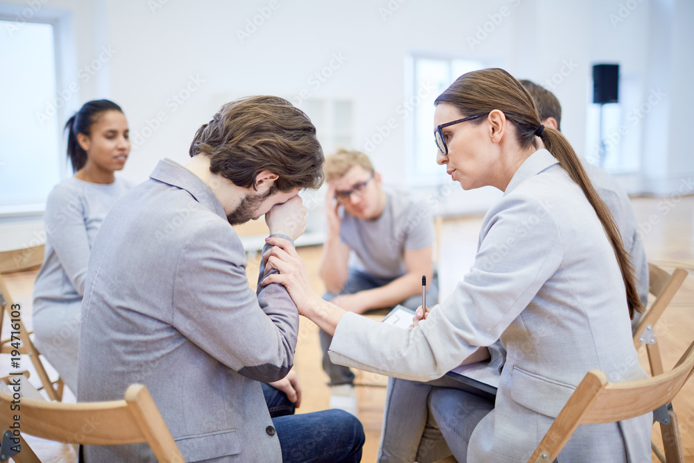 Crying businessman sitting on chair among his colleagues while psychologist reassuring him Crying businessman sitting on chair among his colleagues while psychologist reassuring him