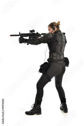 full length portrait of female soldier wearing black tactical armour, standing with back to the camera holding a gun, isolated on white studio background.