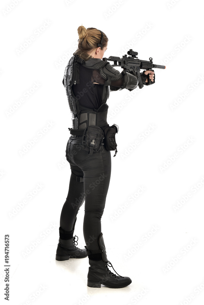 full length portrait of female soldier wearing black tactical armour, standing with back to the camera holding a gun, isolated on white studio background. full length portrait of female soldier wearing black tactical armour, standing with back to the camera holding a gun, isolated on white studio background.