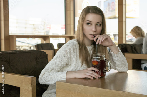 Beautiful young teenage caucasian woman in sweater sitting at the cafe thinking a bit sad in spring (autumn) cold windy weather. lifestyle portrait in nature. Copy space