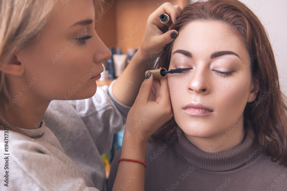 Professional make up artist apply mascara to a young caucasian woman on her date and wedding photosession. Indoors, studio. Casual outfit. Backstage of a process