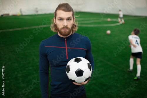 Fototapeta Naklejka Na Ścianę i Meble -  Young muscular sports trainer in activewear looking at camera while his team training on pitch