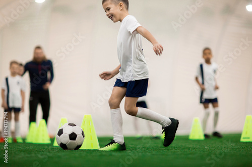 Happy boy in uniform kicking the ball while running around cones during training © pressmaster
