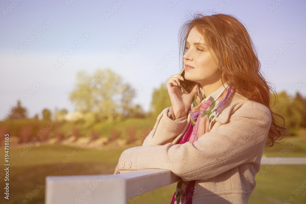 Cute ordinary caucasian brunette woman in casual outfit talking on a phone working while walking at the nature park. Cold spring (autumn) weather, warm coat on a girl. Sunset. Copy space