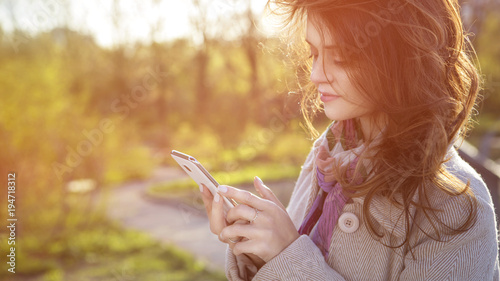 Cute ordinary caucasian brunette woman in casual outfit talking on a phone working while walking at the nature park. Cold spring (autumn) weather, warm coat on a girl. Sunset. Copy space