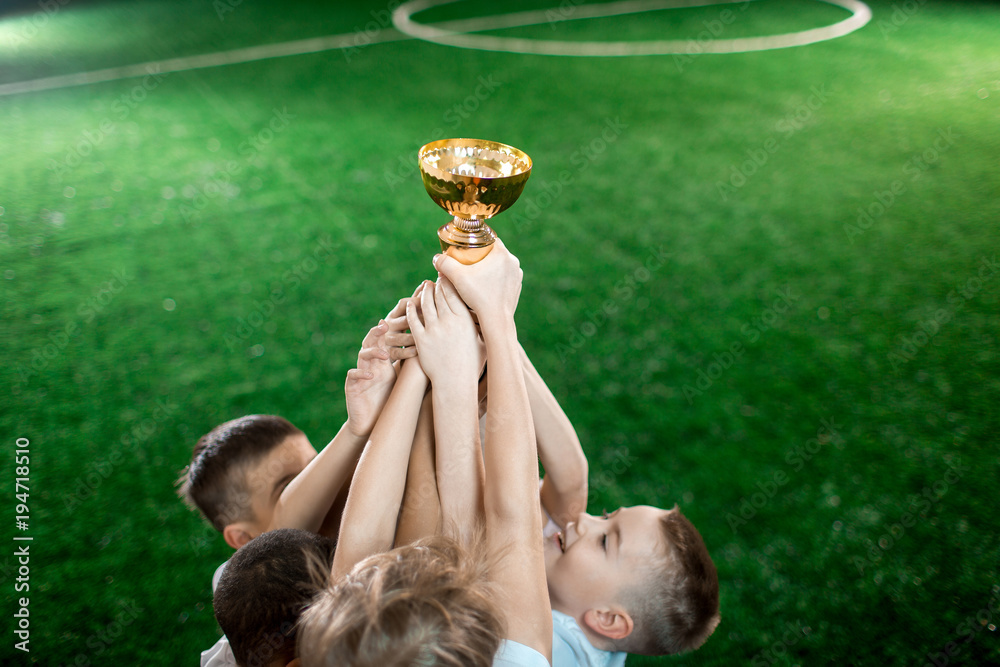 Fototapeta premium Stretched arms of little footballers holding gold award on background of green pitch