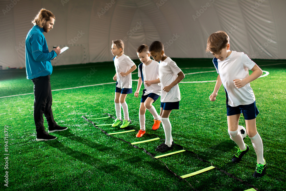 Trainer writing down results of little football players while they exercising on pitch facilities