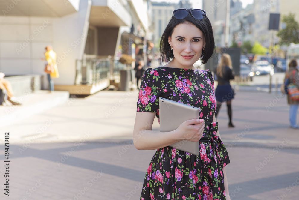 Smart beautiful caucasian fit business woman with a strong face type and dark bob short hairstyle in casual romantic dress in european city streets with tablet in hands, working, smiling. copy space Smart beautiful caucasian fit business woman with a strong face type and dark bob short hairstyle in casual romantic dress in european city streets with tablet in hands, working, smiling. copy space
