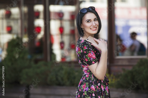 Smart beautiful caucasian fit business woman with a strong face type and dark bob short hairstyle in casual romantic dress in european city streets with phone in hands, working, smiling. copy space © AnnaDemy