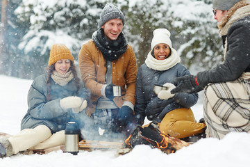 Joyful young people in winterwear sitting in snow arounf fire and having hot drinks