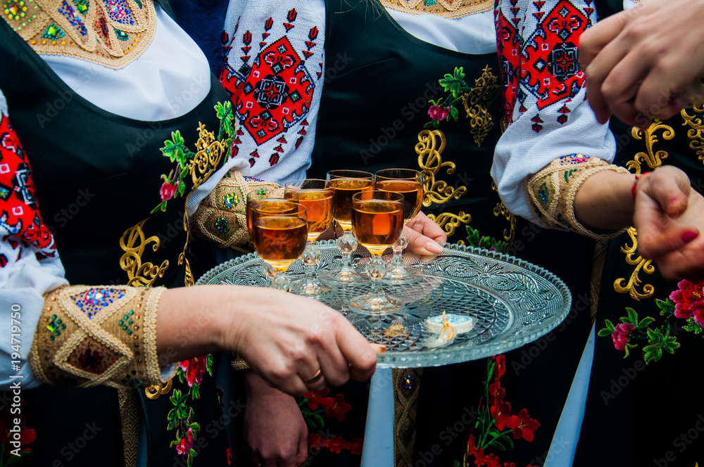Women in traditional ethnic Ukrainian  costumes with patterns holding a silver tray with glasses of wine or cognac - obrazy, fototapety, plakaty 