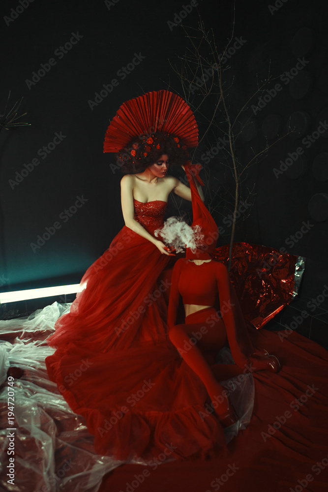 Beautiful model wearing red dress and crown is posing in a dark surreal studio