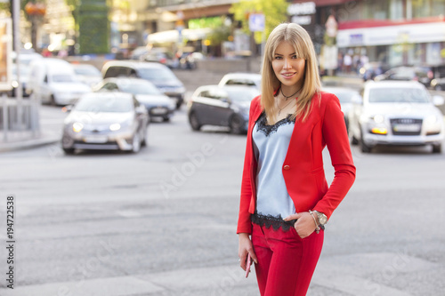 Beautiful rich luxury looking blonde caucasian business woman in smart-casual red outfit standing on a european streets near cars, holding tablet, distant working. Daylight, outdoors.