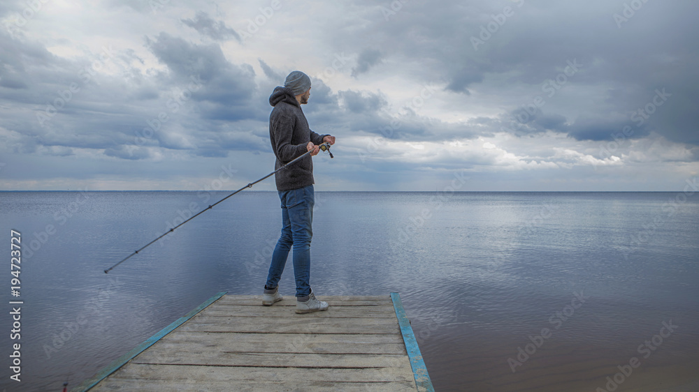 Young handsome brutal caucasian man in casual outfit fishing on a lake as a hobby, trying to catch carp on spinning rod alone. Summer weather, sunset, natural landscapes. HDR toned image