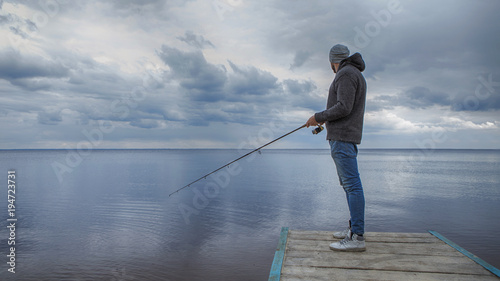 Young handsome brutal caucasian man in casual outfit fishing on a lake as a hobby, trying to catch carp on spinning rod alone. Summer weather, sunset, natural landscapes. HDR toned image