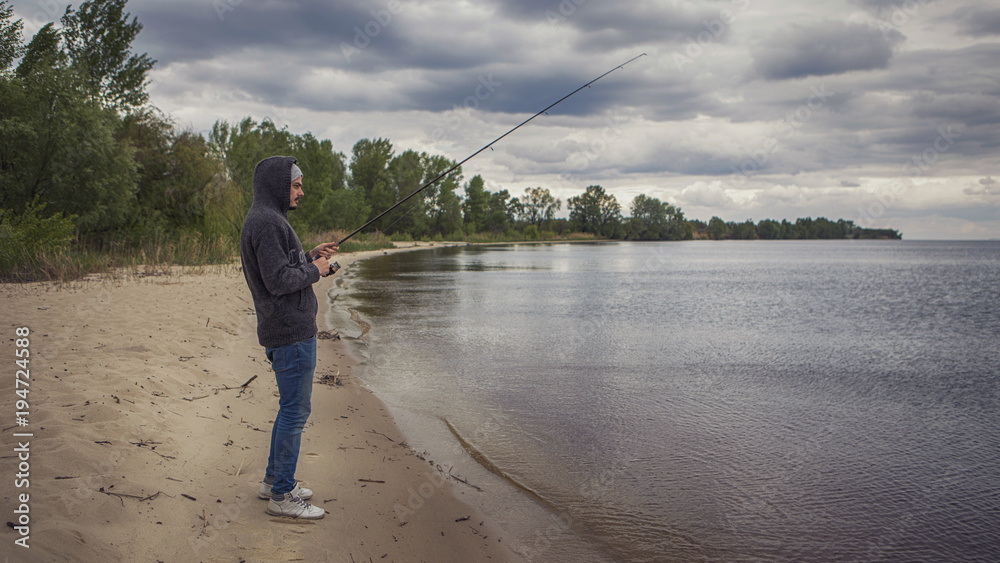 Young handsome brutal caucasian man in casual outfit fishing on a lake as a hobby, trying to catch carp on spinning rod alone. Summer weather, sunset, natural landscapes. HDR toned image Young handsome brutal caucasian man in casual outfit fishing on a lake as a hobby, trying to catch carp on spinning rod alone. Summer weather, sunset, natural landscapes. HDR toned image