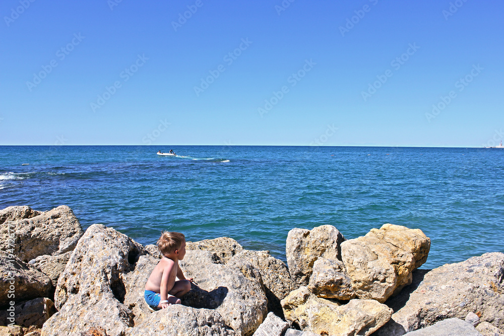 The blue Mediterranean sea in the morning. A little boy is sitting on the rocks on the beach and looking at the sea. Summer vacation.