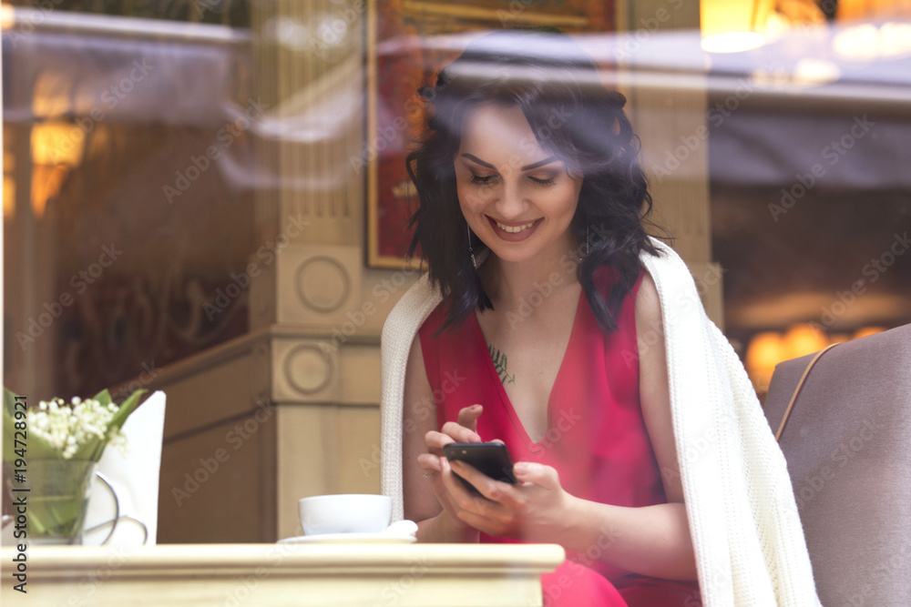 Beautiful brunette caucasian business woman sitting in cafe on a coffee break in red  casual but a bit classic dress and white cardigan, smile, talking on the phone. Copy space. Evening, sunset light