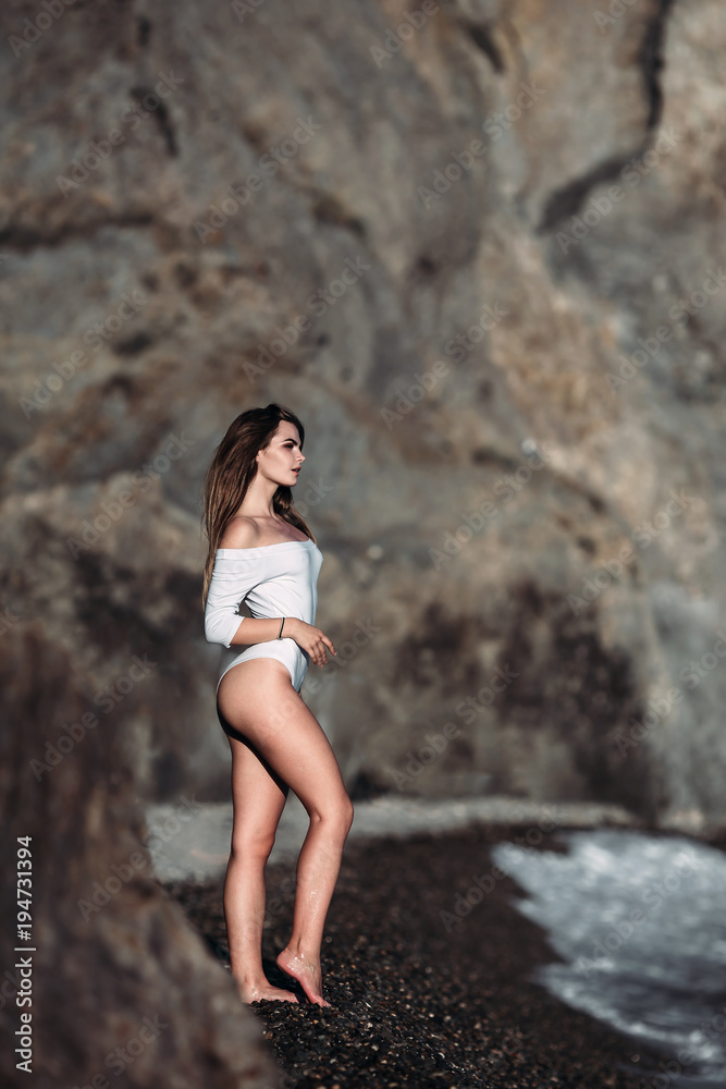 A beautiful girl in a white swimsuit which stands next to a large stone on the rocks, has a magnificent sea view on the beach. Blurred cliff on background. Copy space, vertical