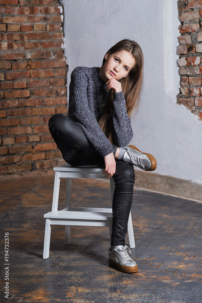 Confident girl sitting in a fashion pose on a chair. Beautiful modern girl posing near the brick wall. Stylish young teenager woman looking at camera raising one's hand to his chin.