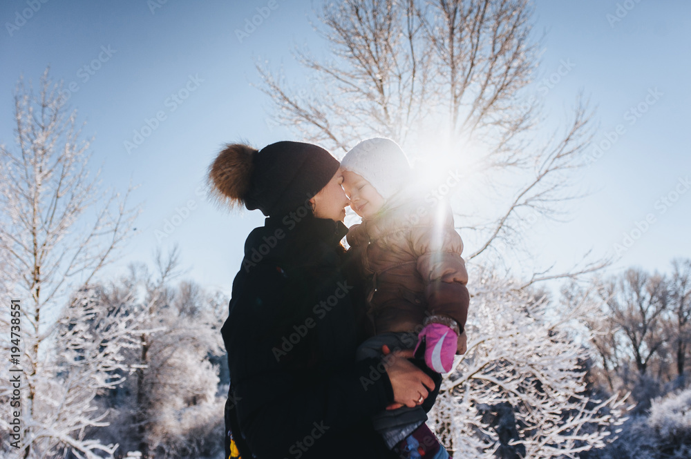 A young mother holds her little daughter in her arms. Winter walk in nature. Mother gently hugs her daughter. Sunlight in winter.
