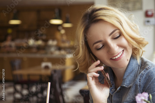 Blonde  with dark roots  caucasian woman in casual summer outfit at the cafe. Grey dress and jeans jacket. Woman got natural day makeup and curly hairstyle. She talks on her phone  working  make calls