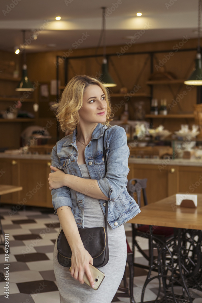 Blonde (with dark roots) caucasian woman in casual summer outfit at the cafe. Grey dress and jeans jacket. Woman got natural day makeup and curly hairstyle. She talks on her phone, working, make calls