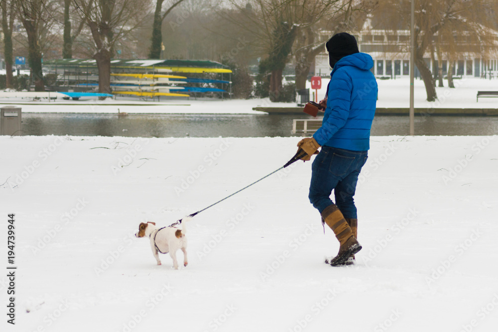 man in blue coat walks jack russell with snow and river in background