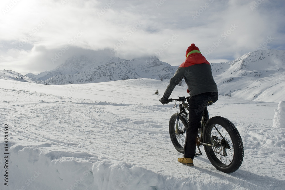man use electric bicycle, e-bike, ebike, pedal on snow covered road, downhill mountain, specific bike with wide wheels to go on snow, called fatbike, winter, cold, alps, Simplon Pass, Switzerland