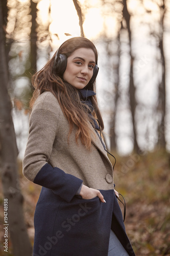 one young woman standing, looking at camera, in forest, wearing winter autumn coat and headphones. side view. © HD92