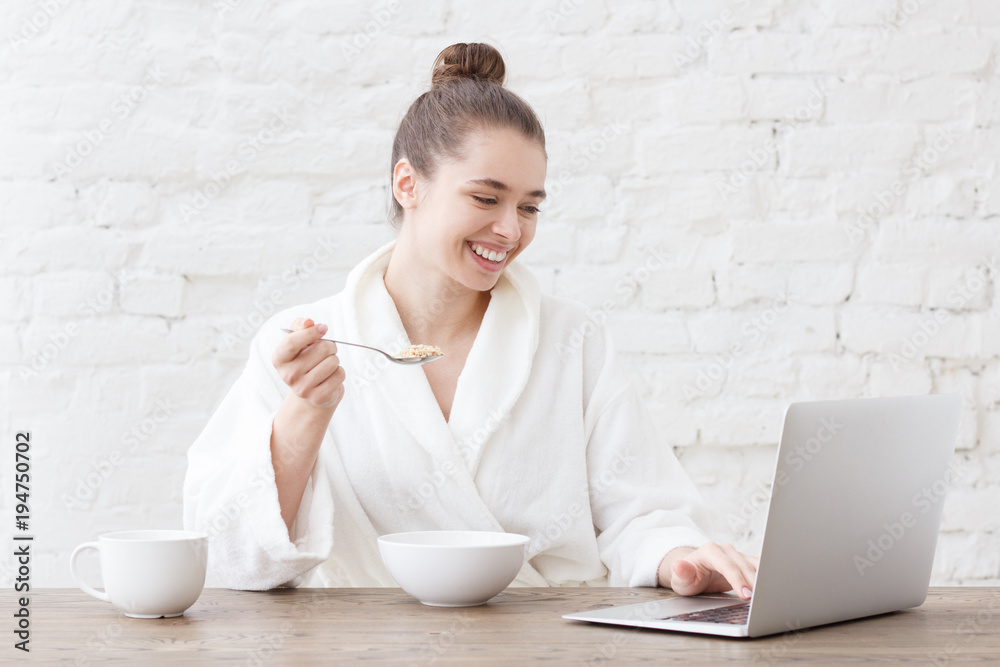 Young cheerful female using her laptop computer while eating breakfast in white loft kitchen Young cheerful female using her laptop computer while eating breakfast in white loft kitchen