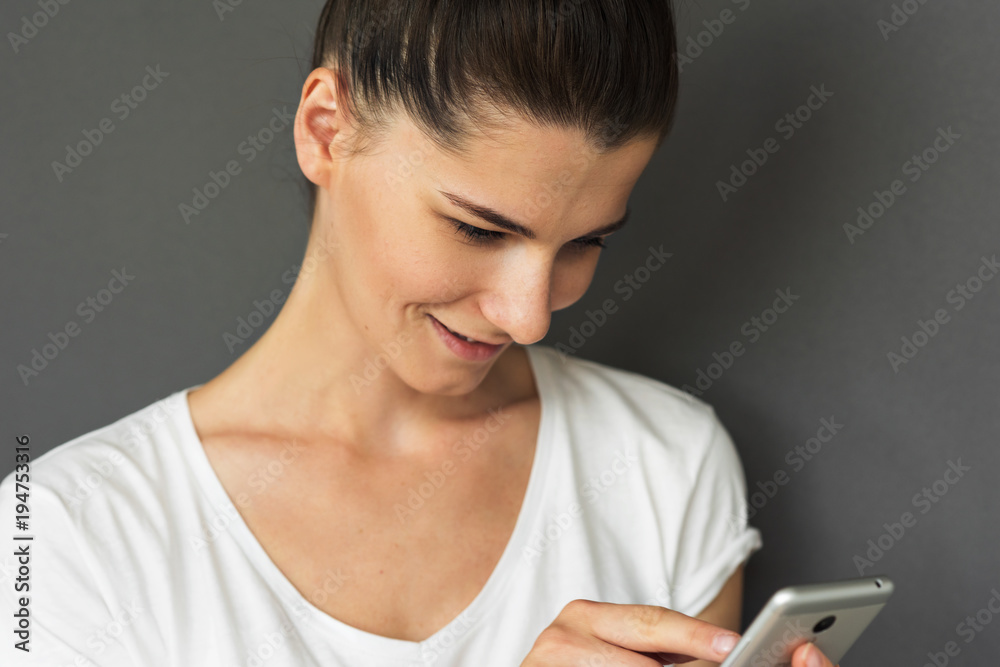 Cropped shot of good looking female in casual whiet T-shirt, smiles happily, reads good news on mobile phone, being satisfied with high speed internet connection. Young woman use social networks. Cropped shot of good looking female in casual whiet T-shirt, smiles happily, reads good news on mobile phone, being satisfied with high speed internet connection. Young woman use social networks.