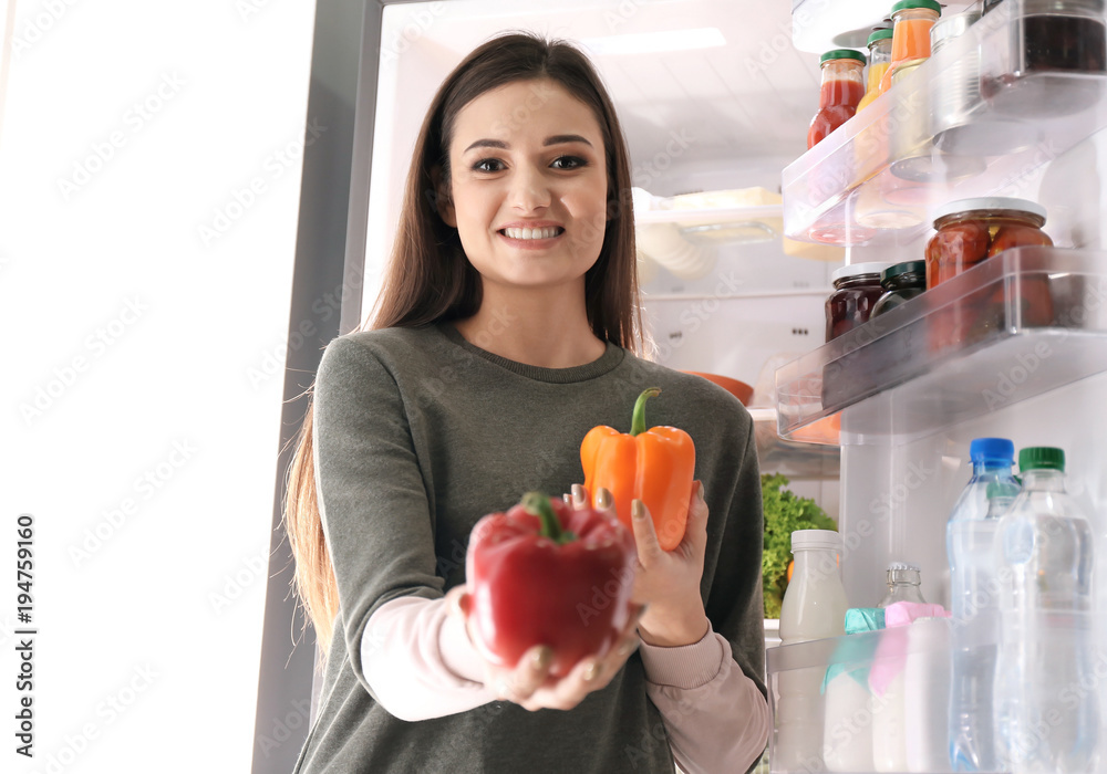 Young woman with ripe bell peppers near open refrigerator at home Young woman with ripe bell peppers near open refrigerator at home