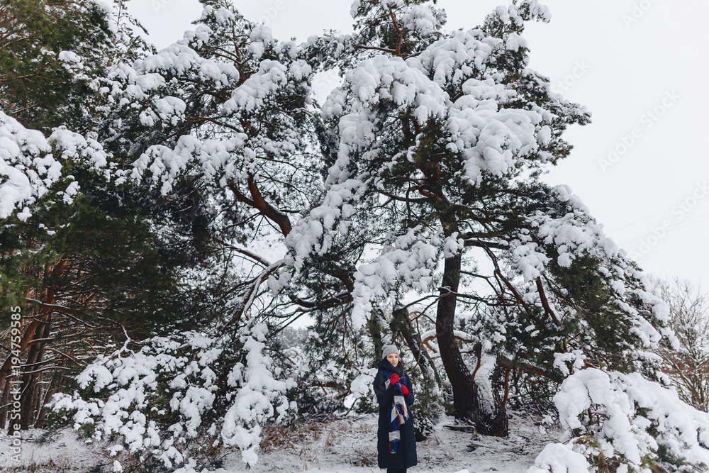 A young, pretty girl poses under a large snow-covered pine forest