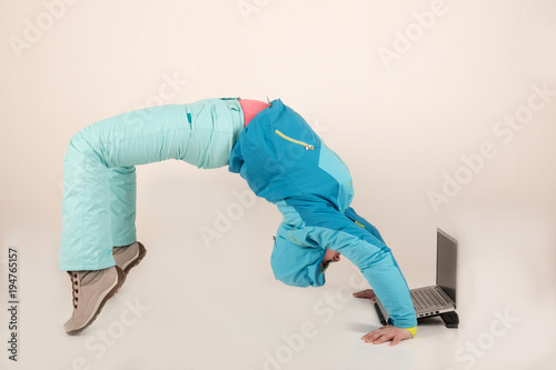 Slender young girl in blue ski suit makes gymnastic exercises in front of a laptop. The concept of sport lifestyle. Busy schedule of young business woman. Sportswear and new computer technologies