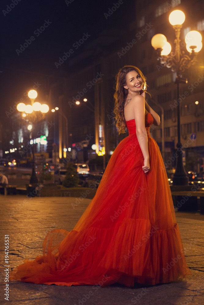 Beautiful sexy caucasian woman with long curly brunette hair and make up on big eyes and full sensual lips. She coming back from party, walking in evening dress on european street with traffic lights