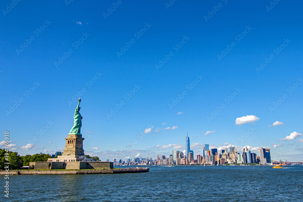 Statue of Liberty in foreground with unmistakeable New York’s Manhattan cityscape in background viewed across the Hudson and against a bright blue sky