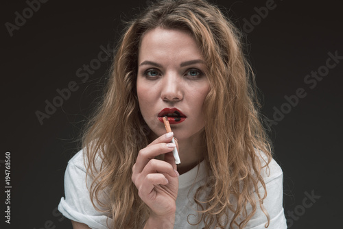 Portrait of discontented blonde woman with wound on her lips, she is holding cigarette and looking at camera. Isolated on background