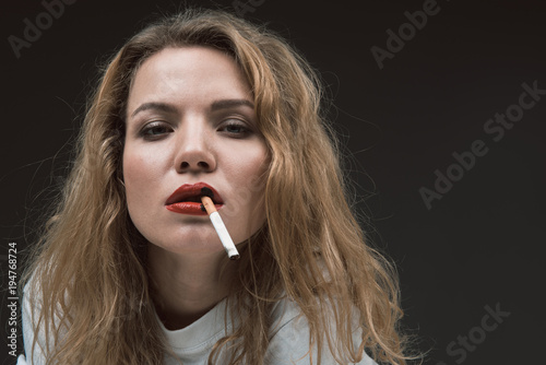 Portrait of beautiful calm blonde girl holding cigarette in mouth, black spots on red lips imitating burned skin. Copy space in right side. Isolated on background